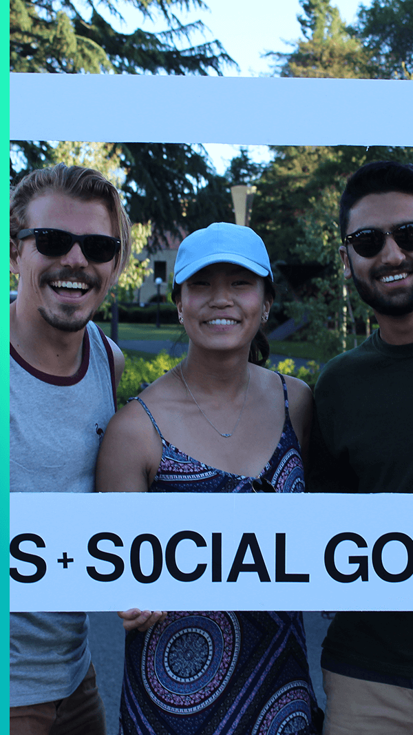 Three smiling students standing on grass holding up a large frame that says CS+Social Good